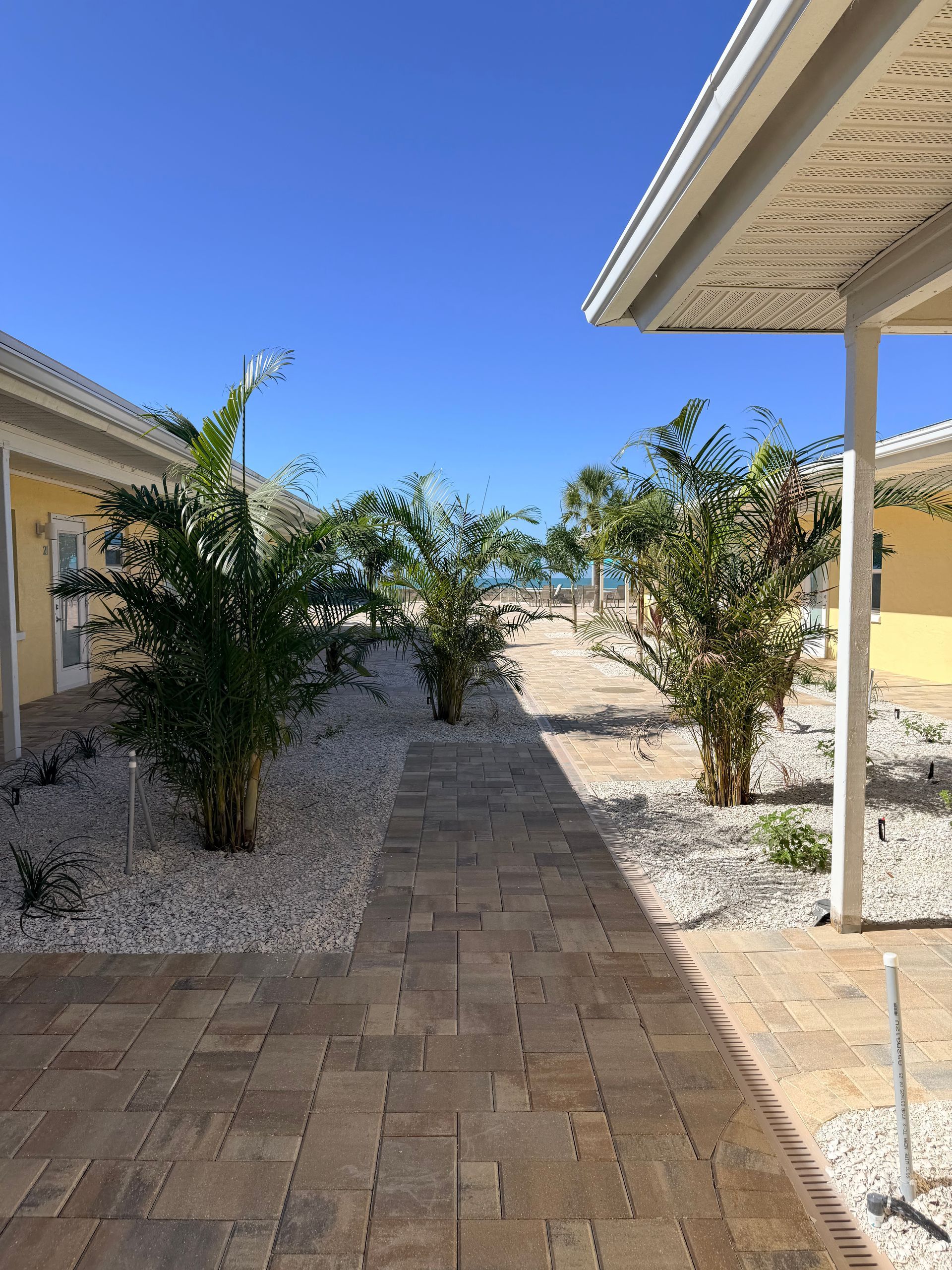 Brick pathway between buildings, dotted with shrubbery, leading to the ocean under a bright blue sky.