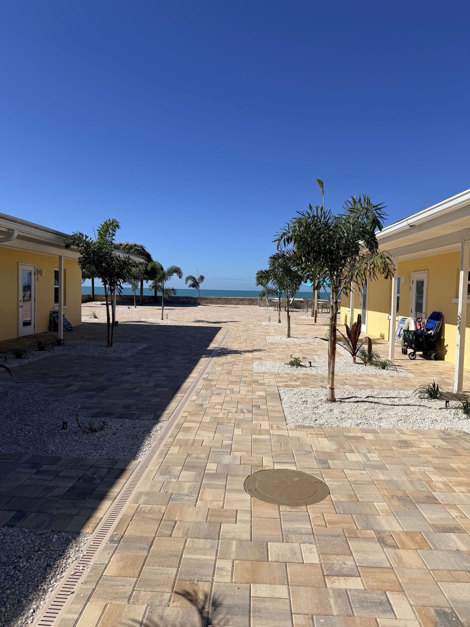 Courtyard with yellow buildings, brick path, small trees, and a view of the ocean under a clear blue sky.