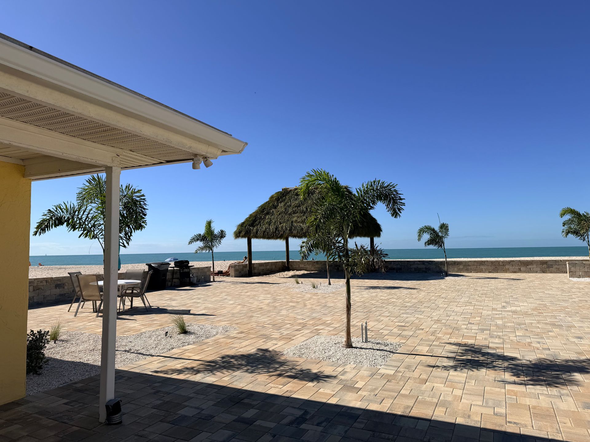 Courtyard with a thatched-roof shelter near the ocean; blue sky, palm trees, and yellow building.