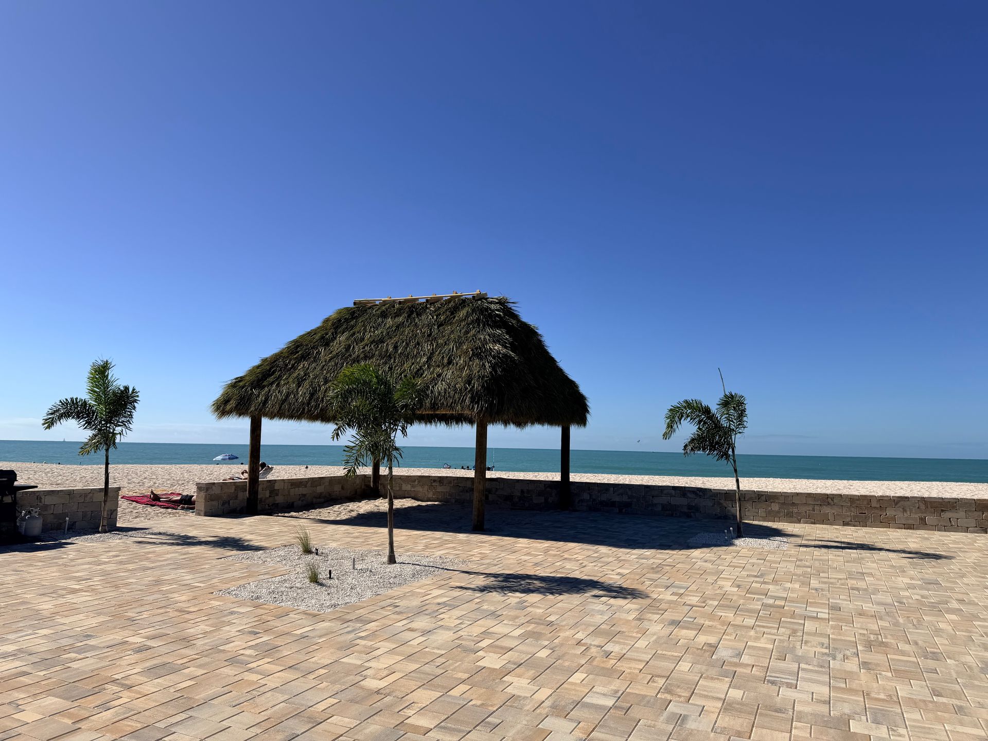 Beachside thatch-roofed shelter on a sunny day. Light-colored bricks, palm trees, and ocean in the background.