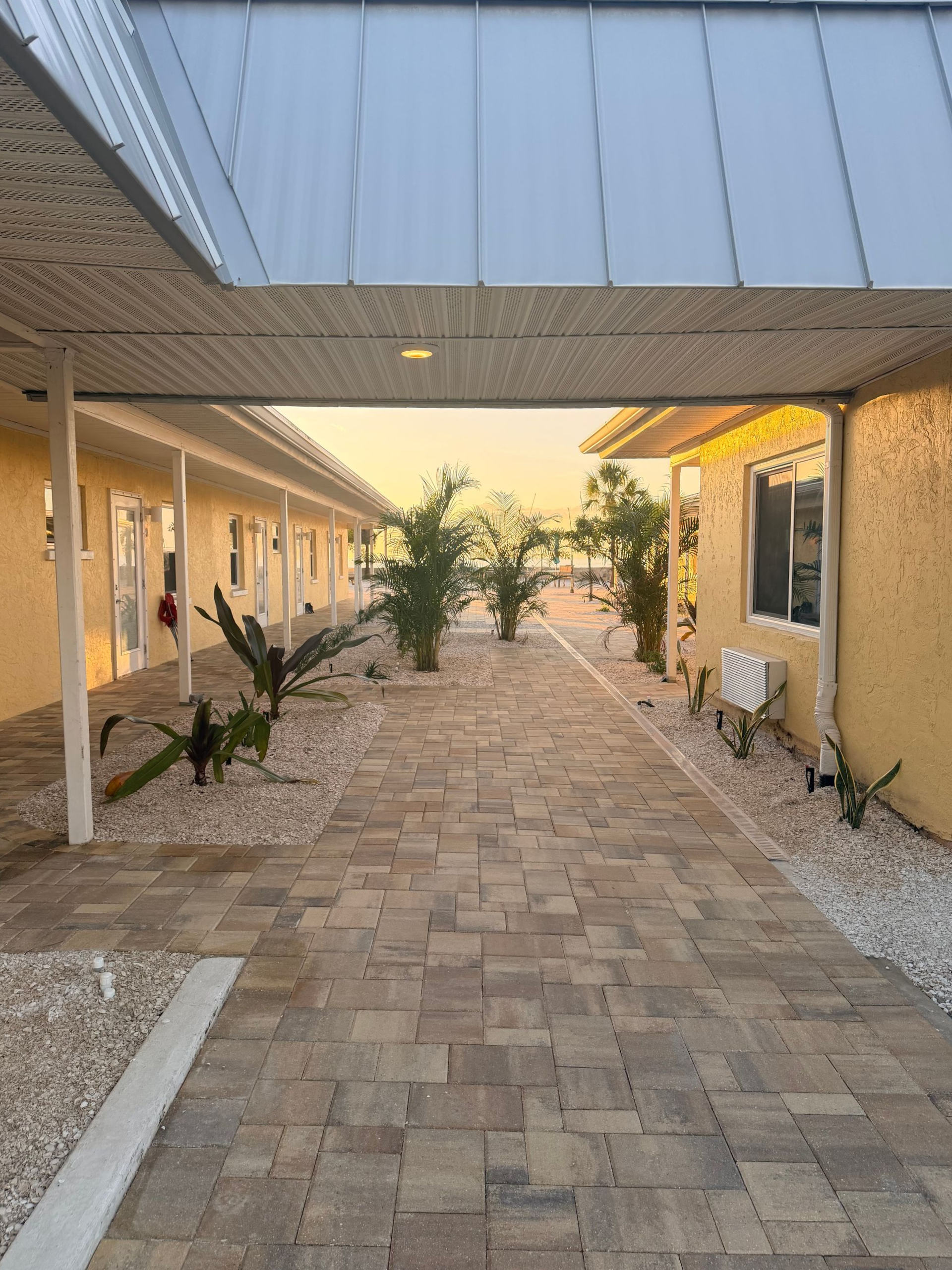 Brick pathway between buildings with a covered walkway. Tropical plants and a sunset are visible.