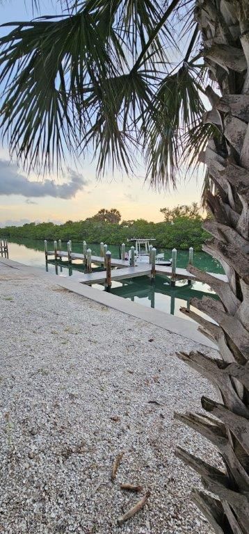 Palm trees framing a small dock over calm water at sunset, with a pebble shore in the foreground