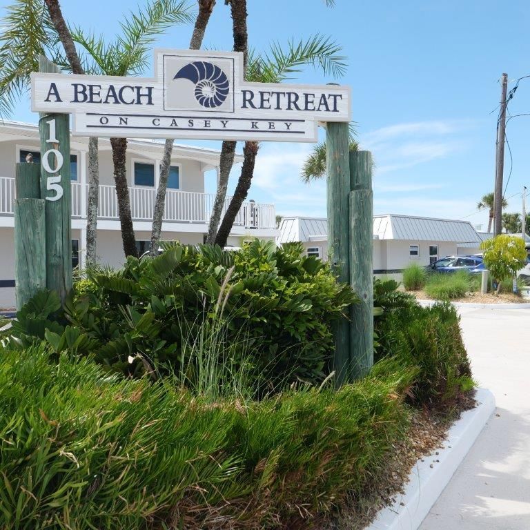 Entrance sign for A Beach Retreat on a sunny coastal road, with palm trees and greenery