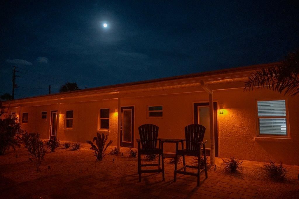 Nighttime view of a lit adobe house with chairs on a patio under a bright moon.