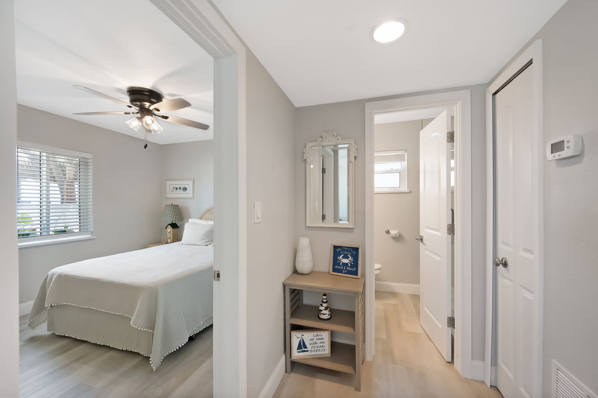Hallway with bedroom on the left and bathroom on the right; neutral walls and light wood floors; decorative mirror and shelf.