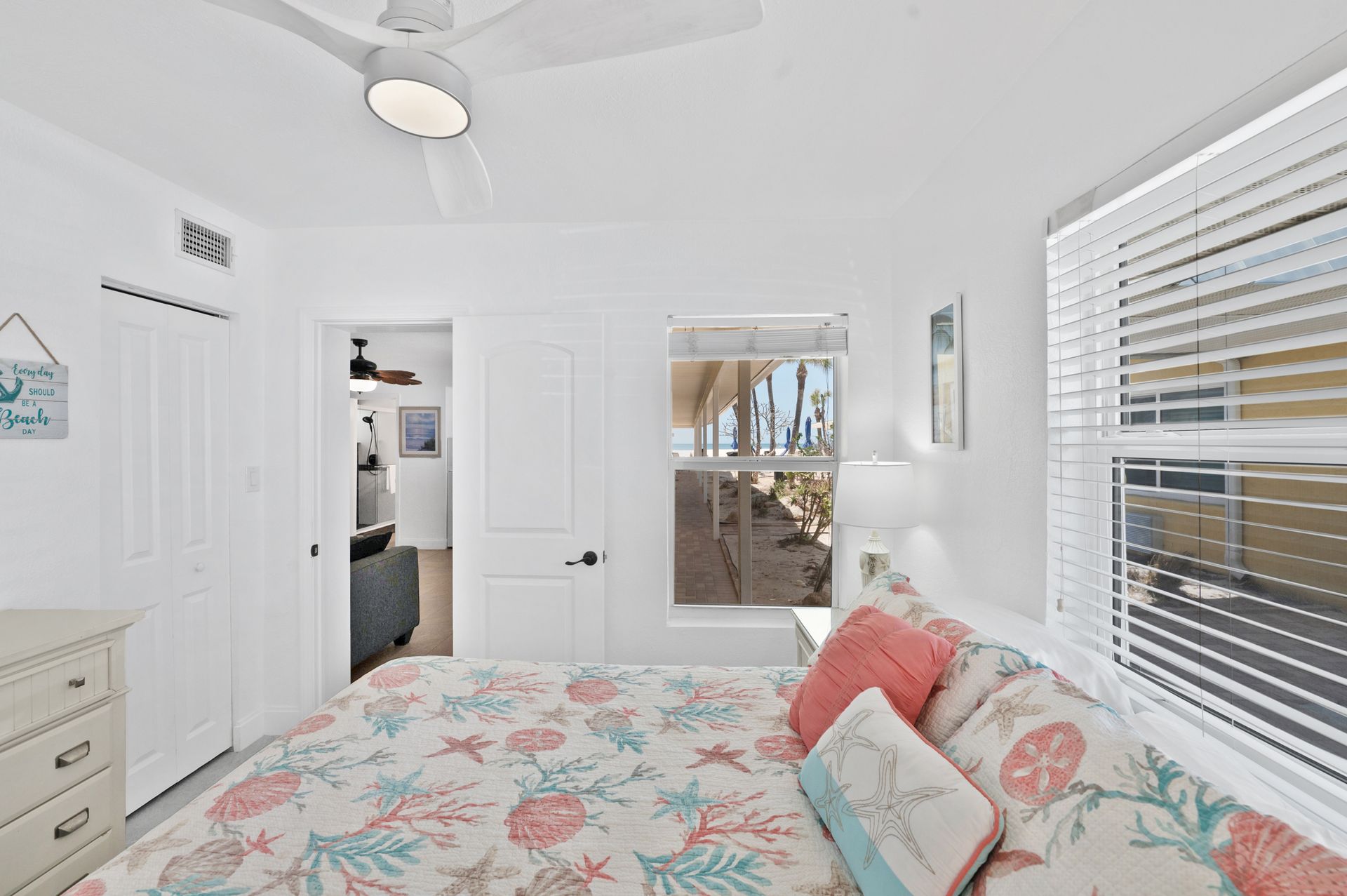 Bedroom with white walls, beach-themed bedding, a window with blinds, and a door leading to another room.
