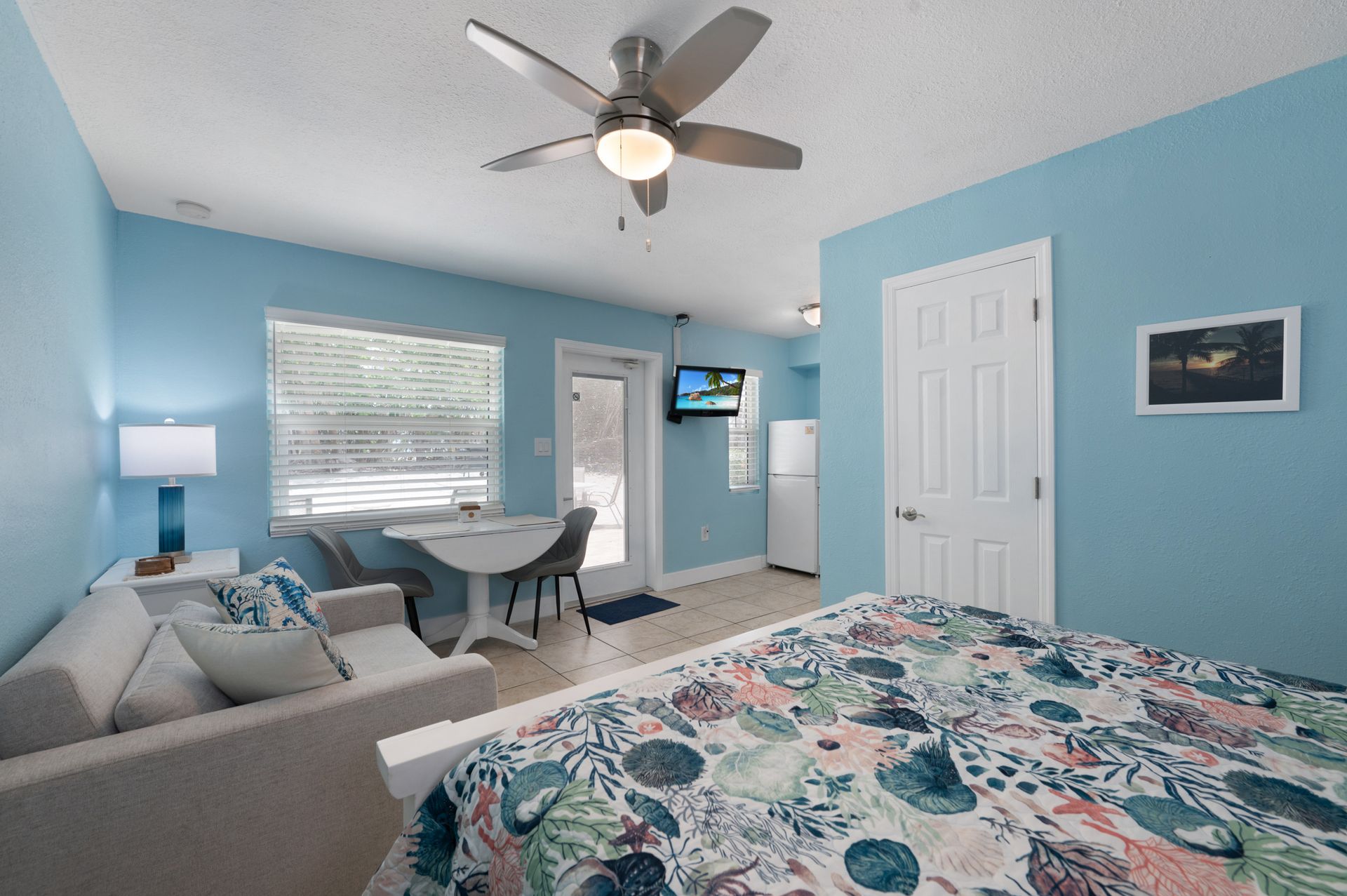 A light blue studio apartment with a bed, small sofa, dining table, and door leading outside.