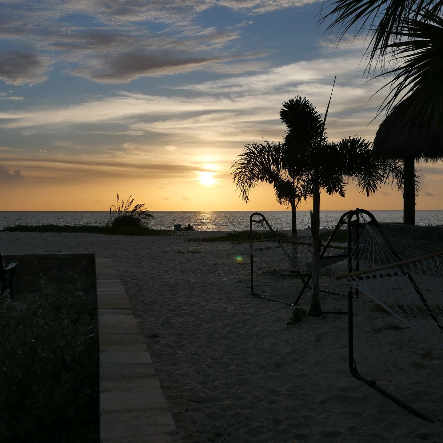 Sunset over a beach with palm trees, sand, and a hammock silhouette by the shore.