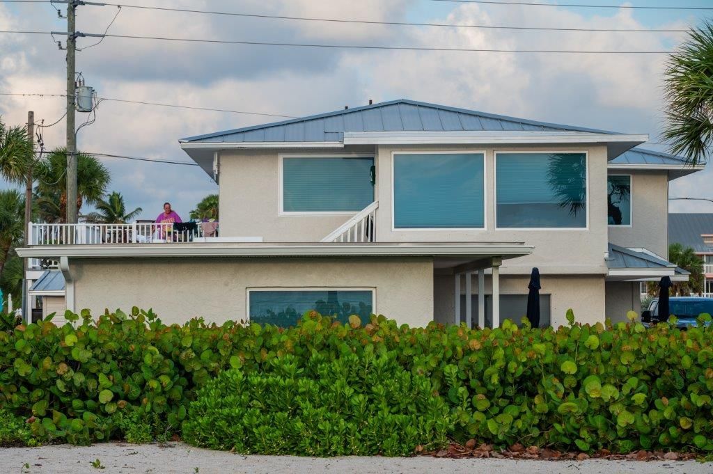 Modern two-story beach house with blue roof and windows, behind a green hedge under a cloudy sky