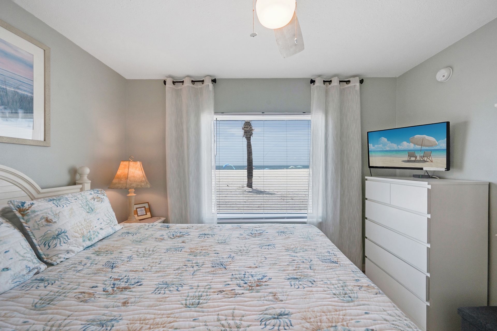 Bright bedroom with floral bedspread, window curtains, bedside lamp, and white dresser with TV.