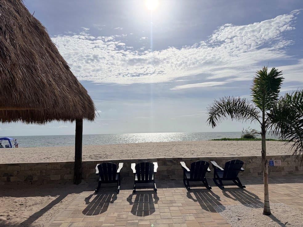Beachfront lounge chairs under thatched shade with ocean horizon and bright sunlit sky