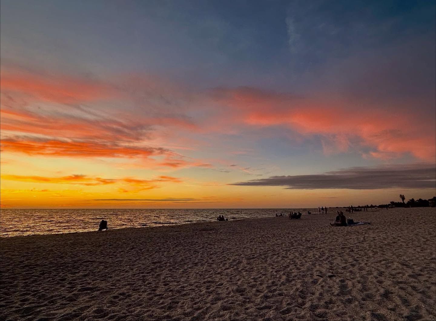 Beach at sunset with orange and pink clouds, people silhouetted on sand.
