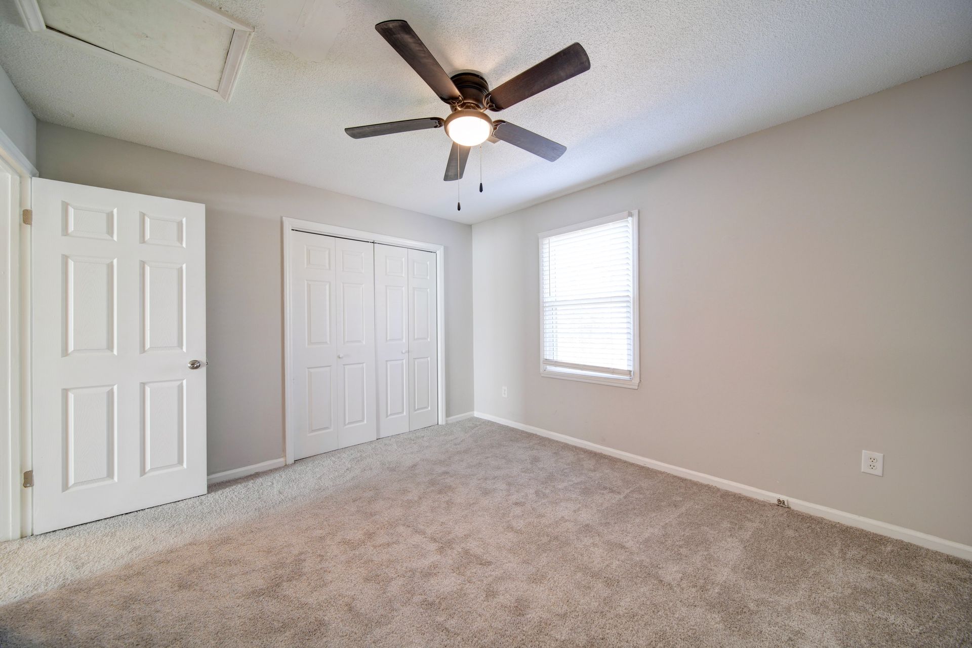 Empty beige bedroom with carpet, ceiling fan, closet doors, and a window
