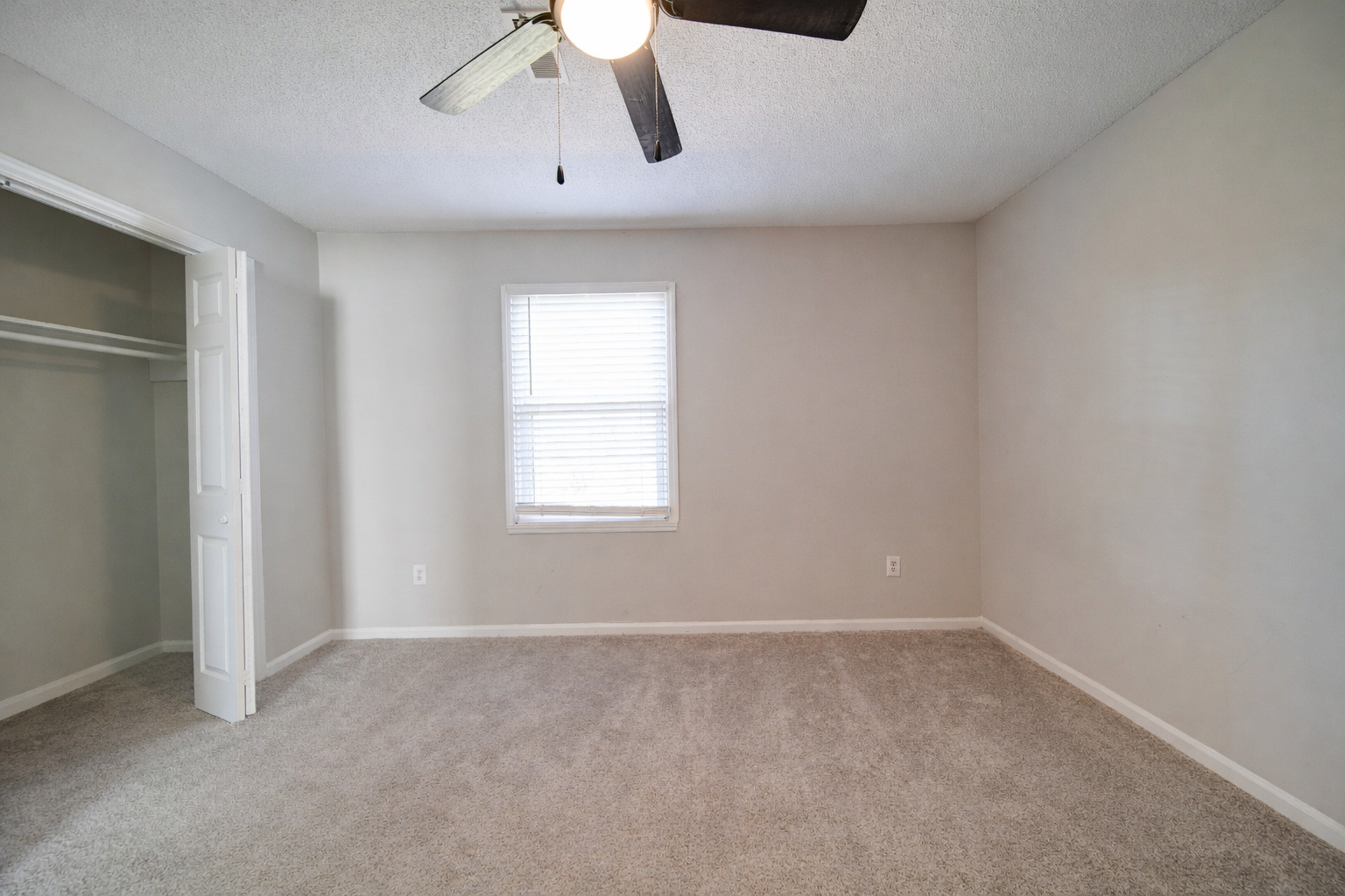 Empty beige bedroom with carpet, window, closet, and ceiling fan