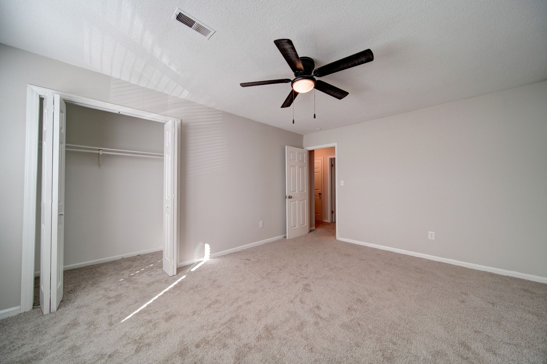 Empty beige bedroom with open closet, carpeted floor, and ceiling fan