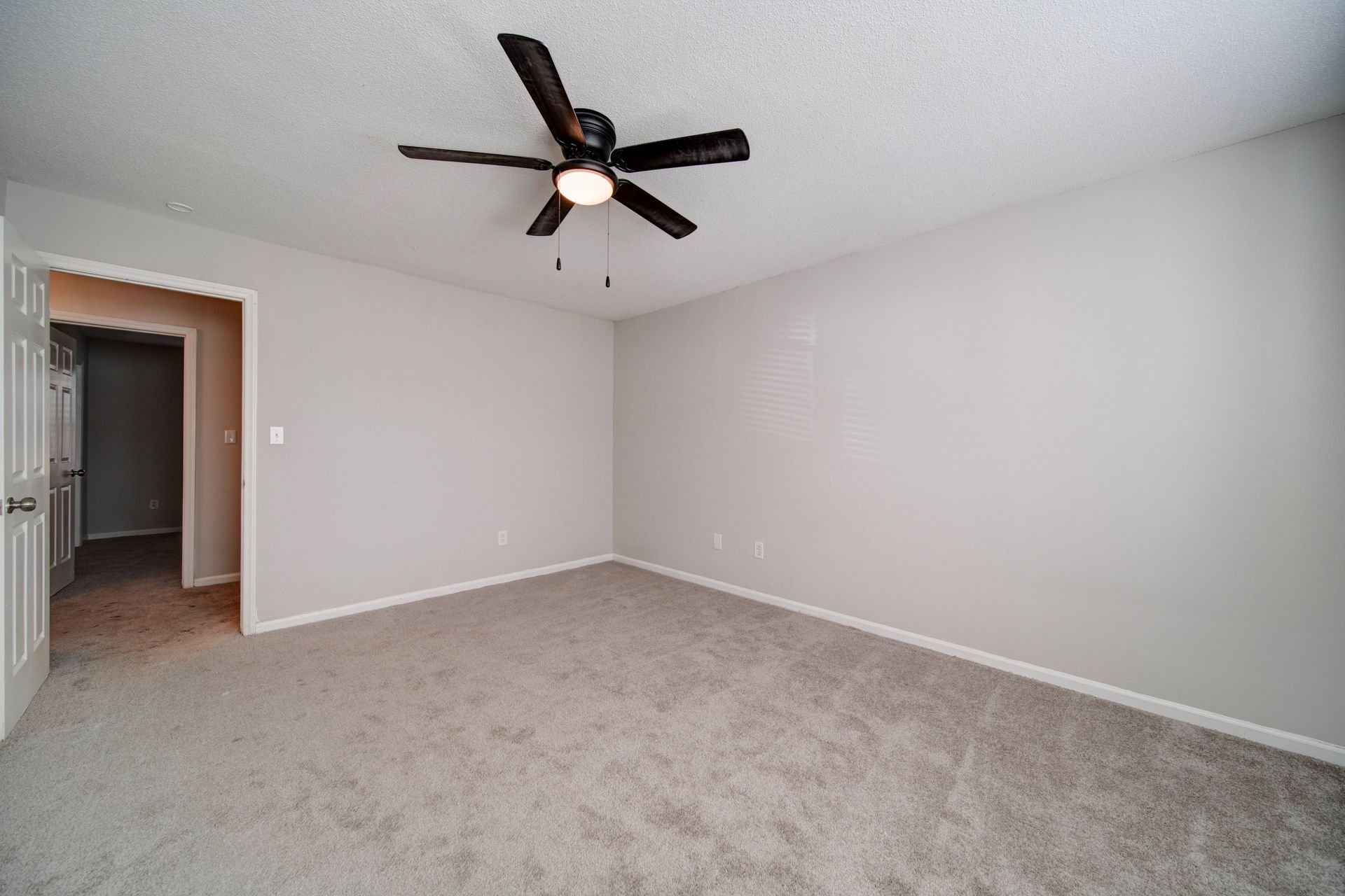 Empty beige bedroom with carpet, ceiling fan, and an open doorway to a hallway