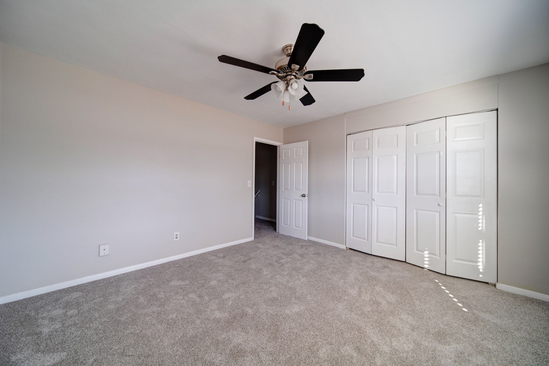 Empty carpeted bedroom with white walls, ceiling fan, and a large closet with sliding doors.