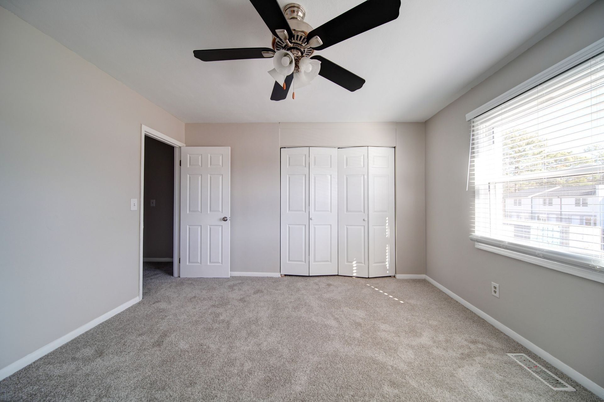 Empty bedroom with gray carpet, white walls, ceiling fan, closet doors, and a window with blinds.