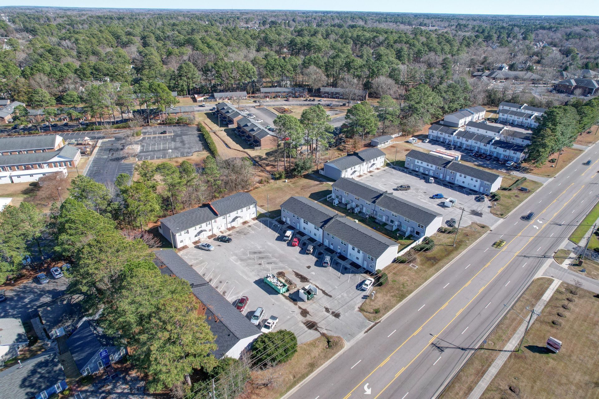 Aerial view of a strip mall and parking lot beside a busy road, surrounded by trees