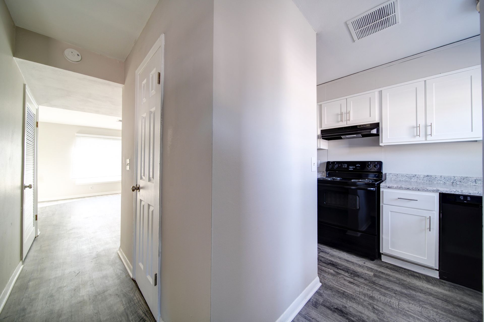 Bright hallway beside a small kitchen with white cabinets, black stove, and gray flooring.