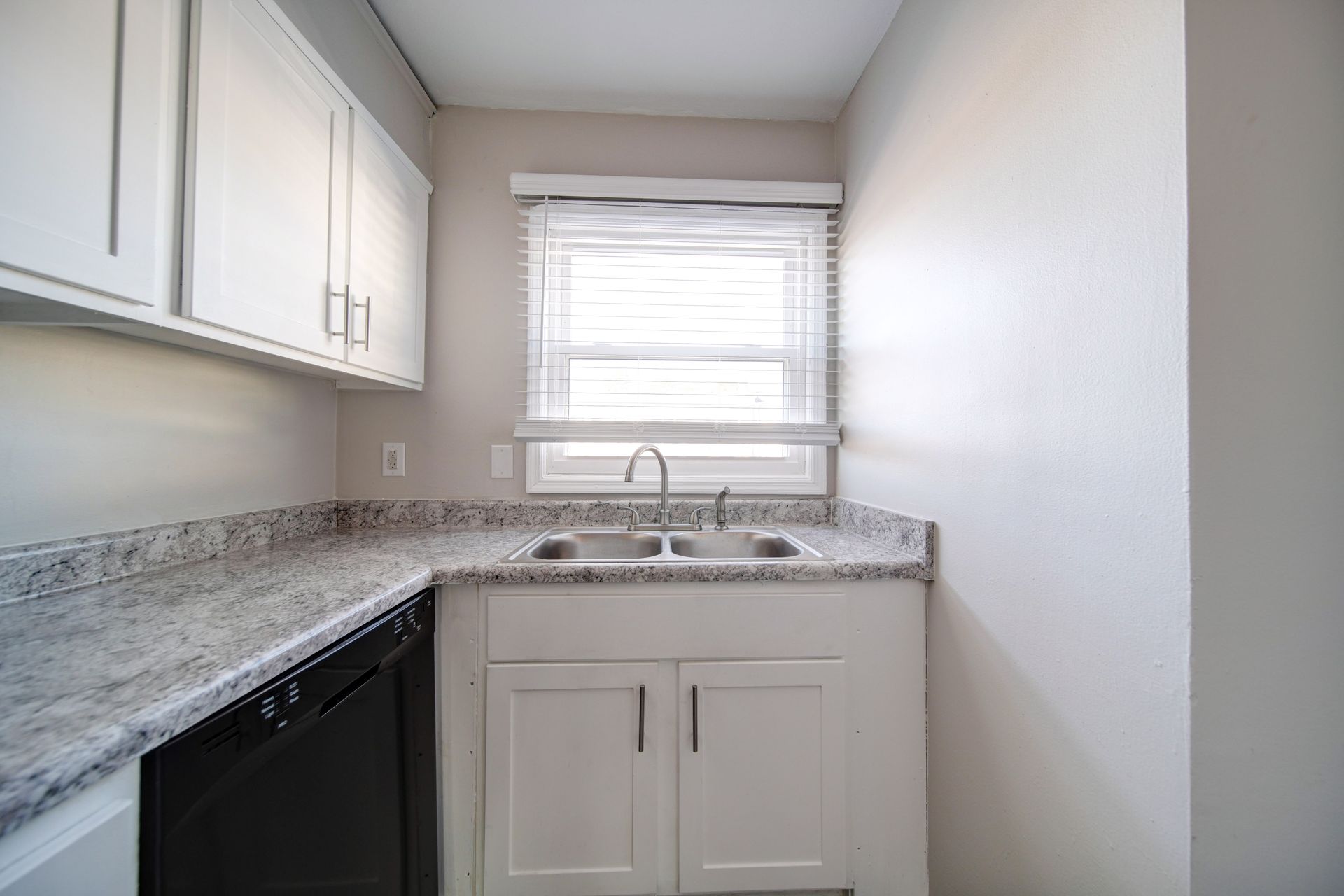 Small kitchen with white cabinets, granite countertops, sink, window, and black dishwasher.