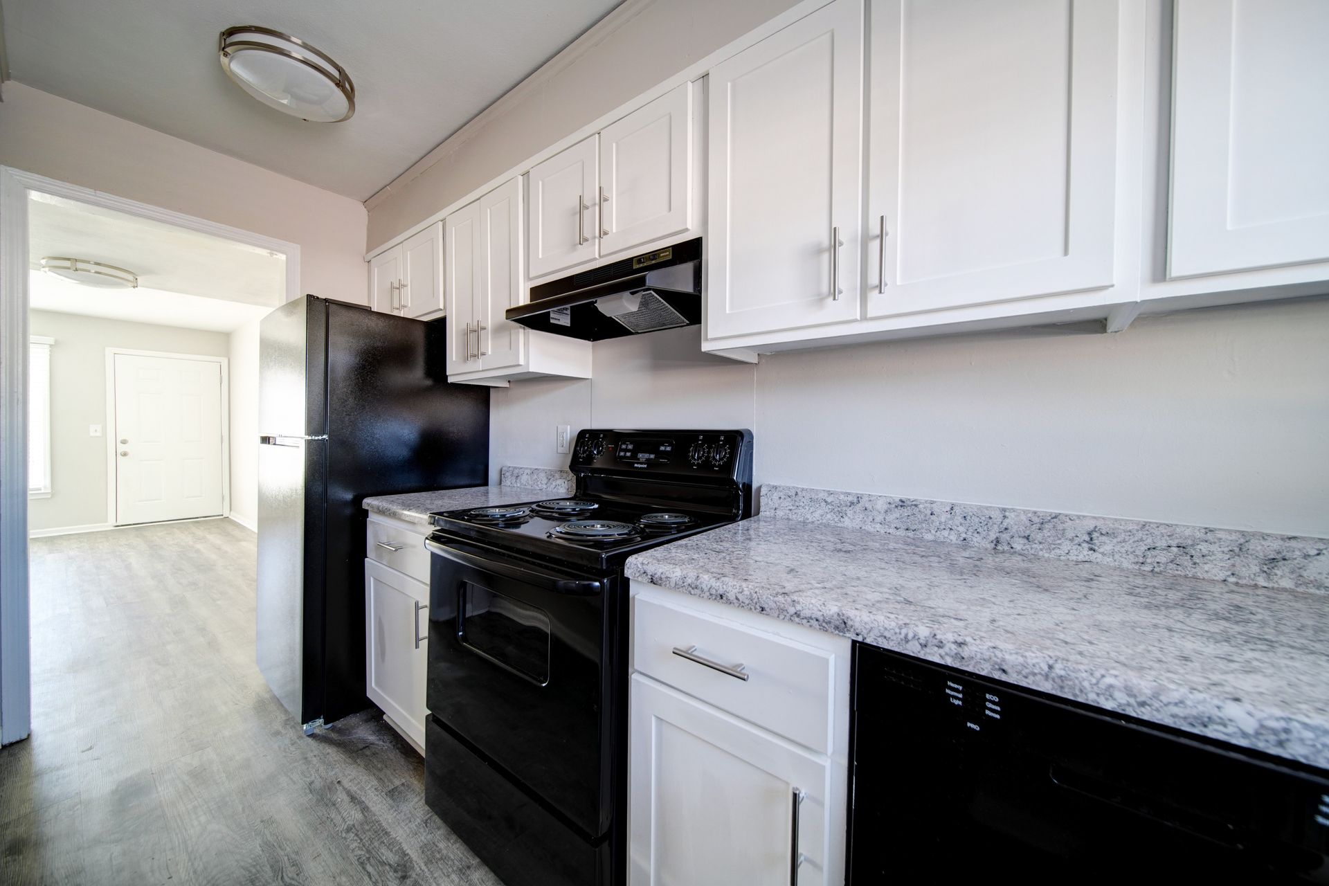 Bright kitchen with white cabinets, black appliances, and speckled countertop, leading into a hallway
