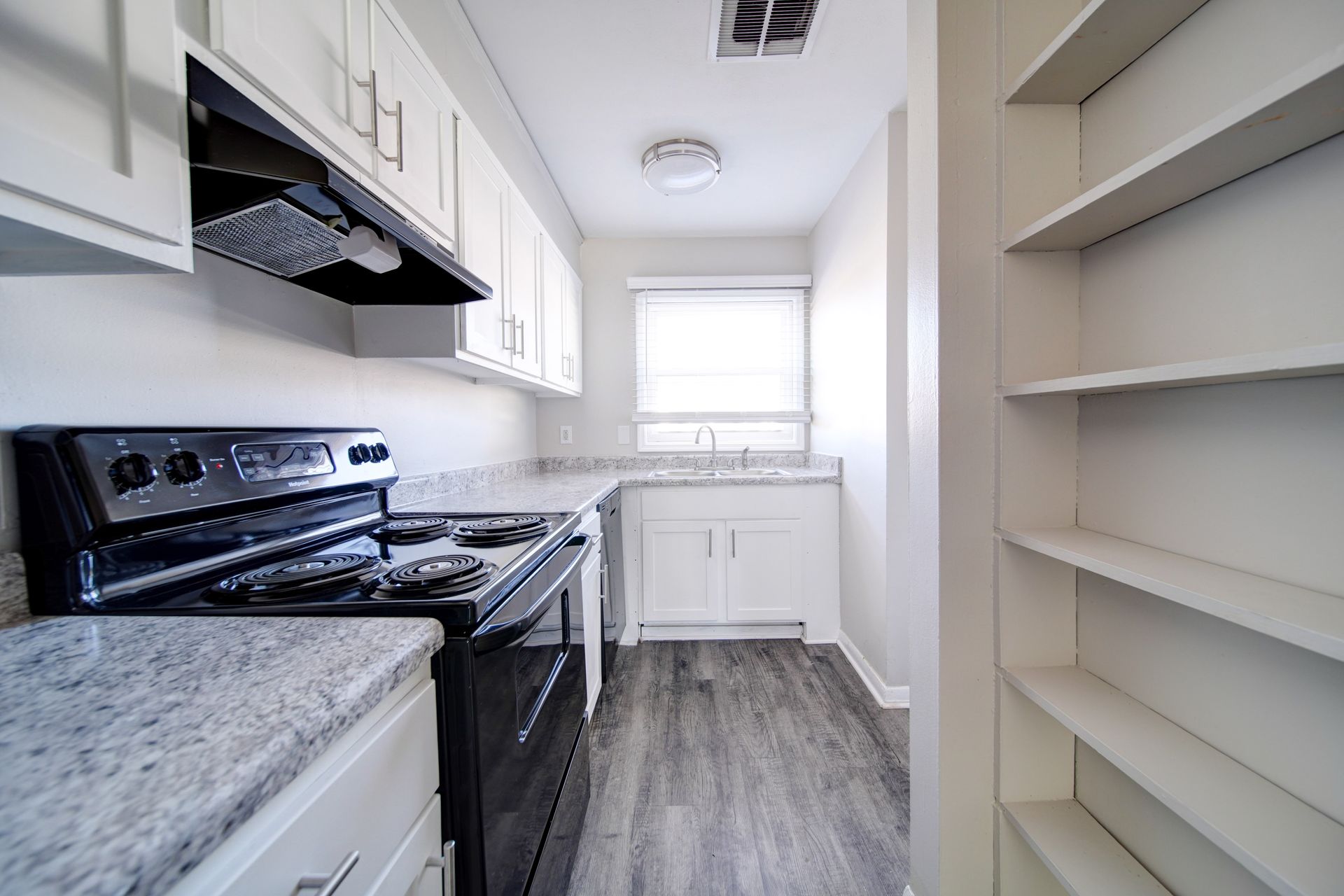 Narrow kitchen with black stove, white cabinets, gray countertops, pantry shelves, and a window at the end.