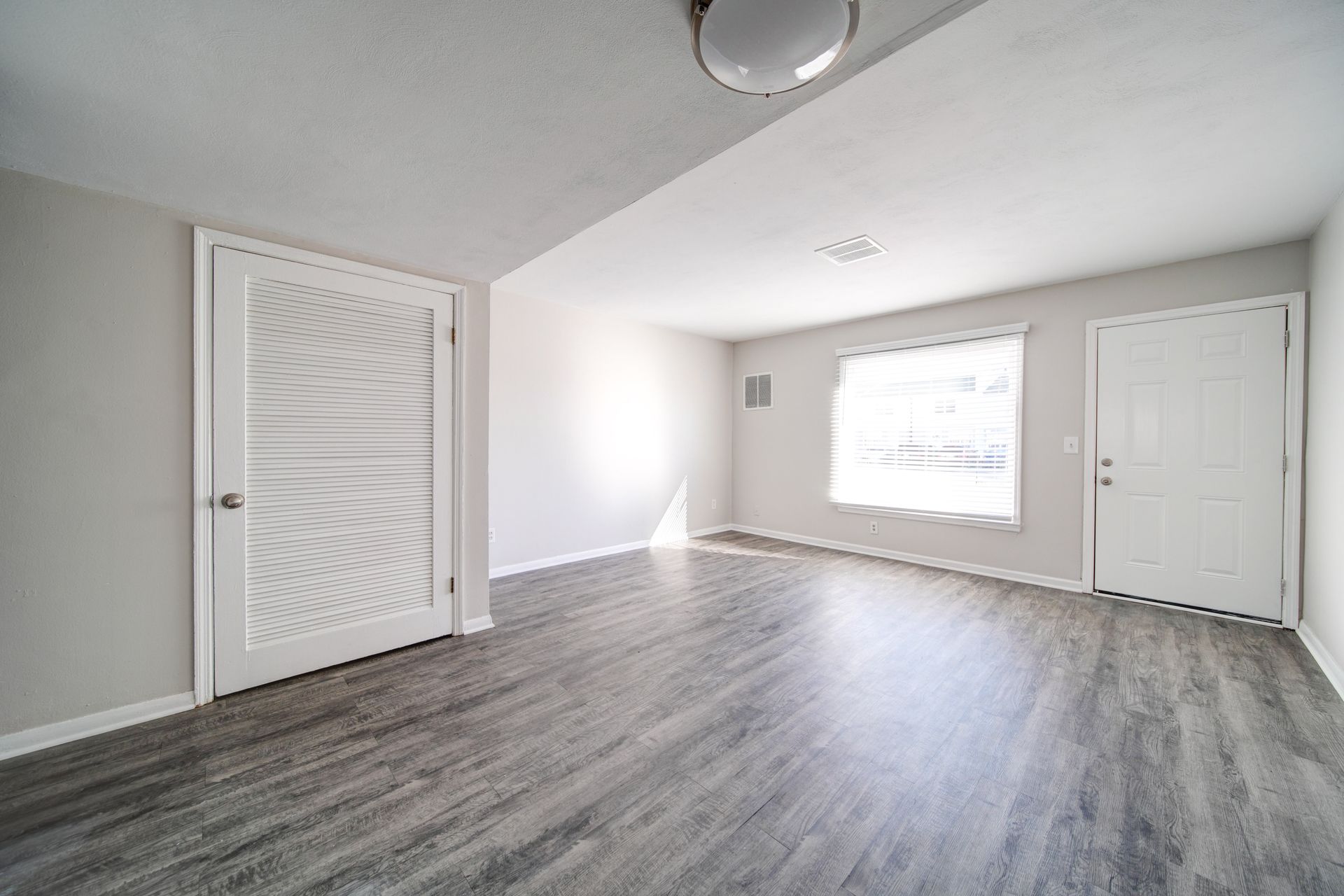 Empty modern room with gray wood floors, white walls, large window, and double closet doors