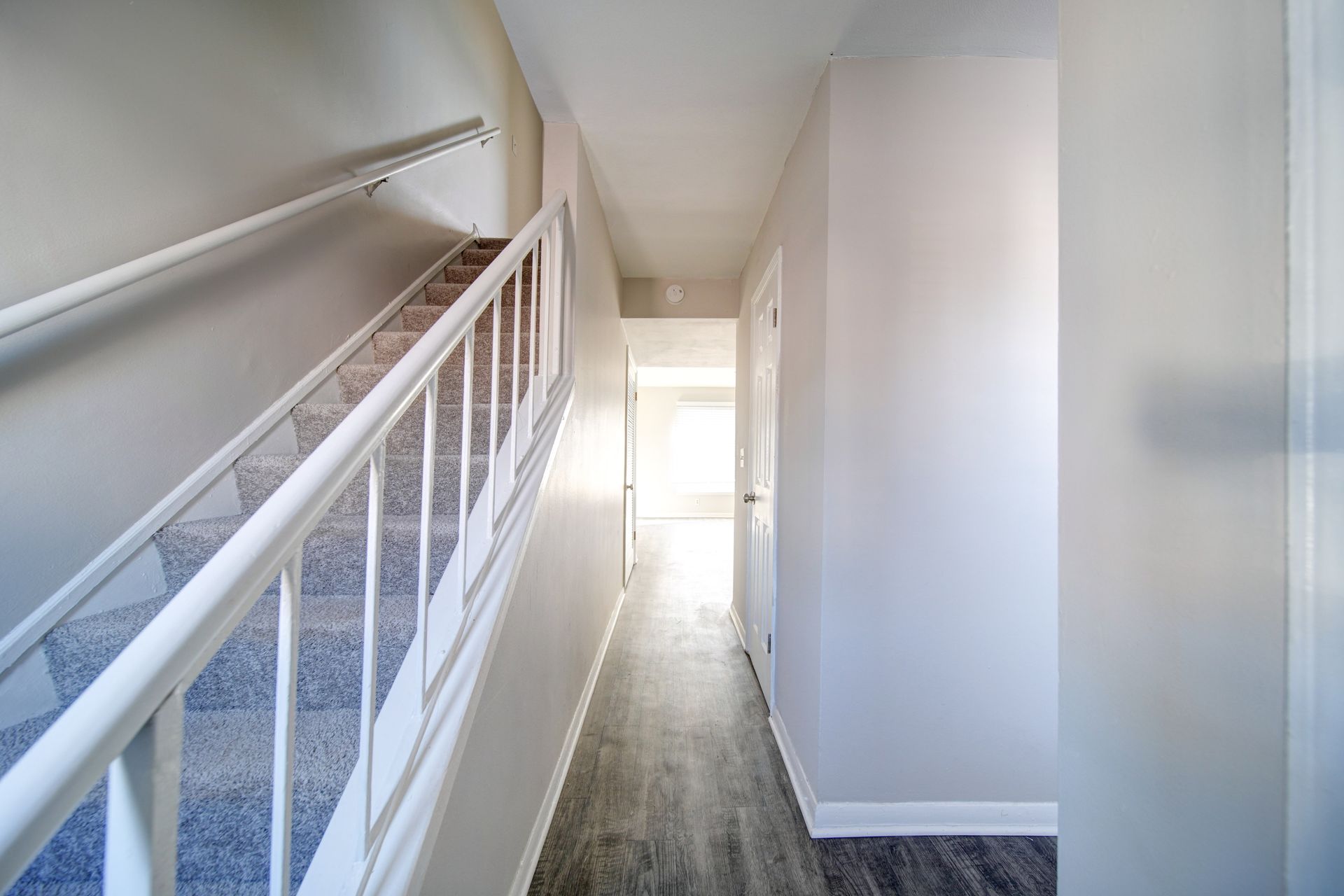 Narrow hallway with white walls, carpeted stairs on the left, and bright light at the far end.