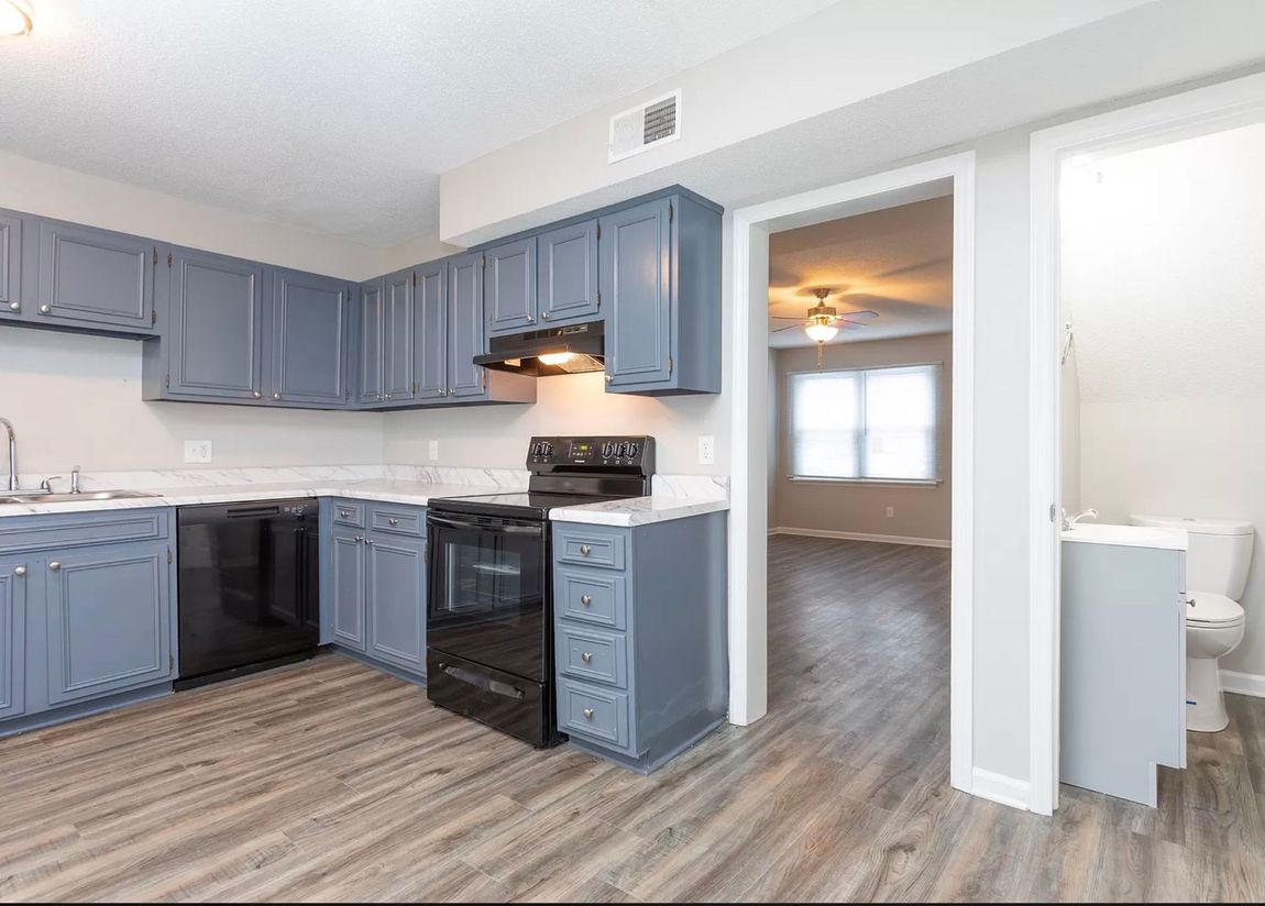 Bright kitchen with blue cabinets, black stove, white walls, and an open doorway to a dining area.