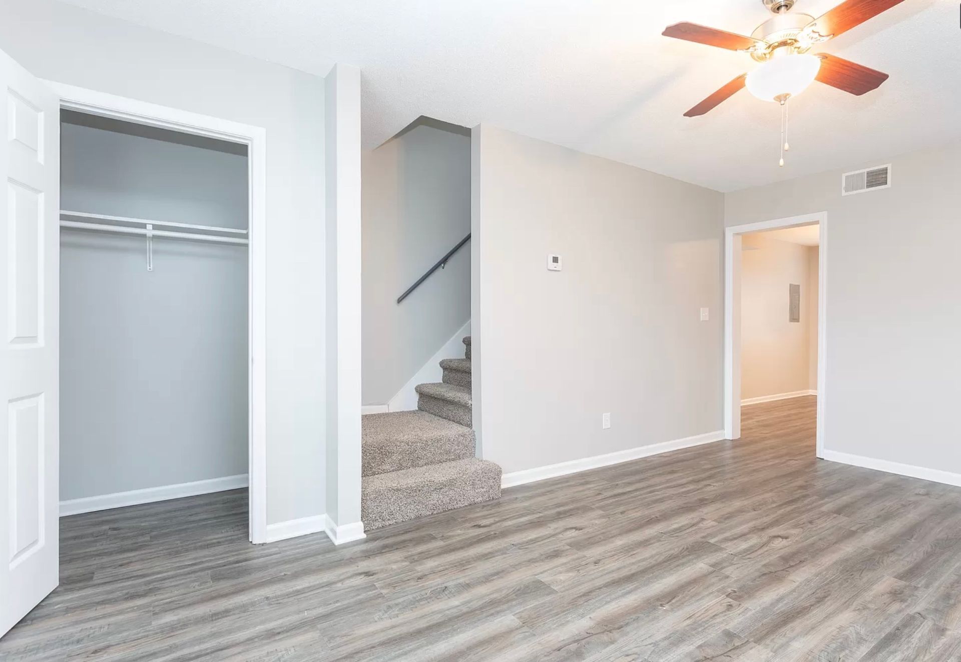 Empty bedroom with gray wood floors, white walls, open closet, and stairs leading up.