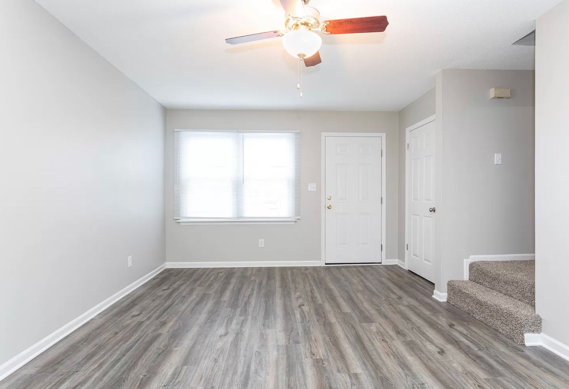 Empty gray living room with wood-look floor, window blinds, ceiling fan, and carpeted stairs