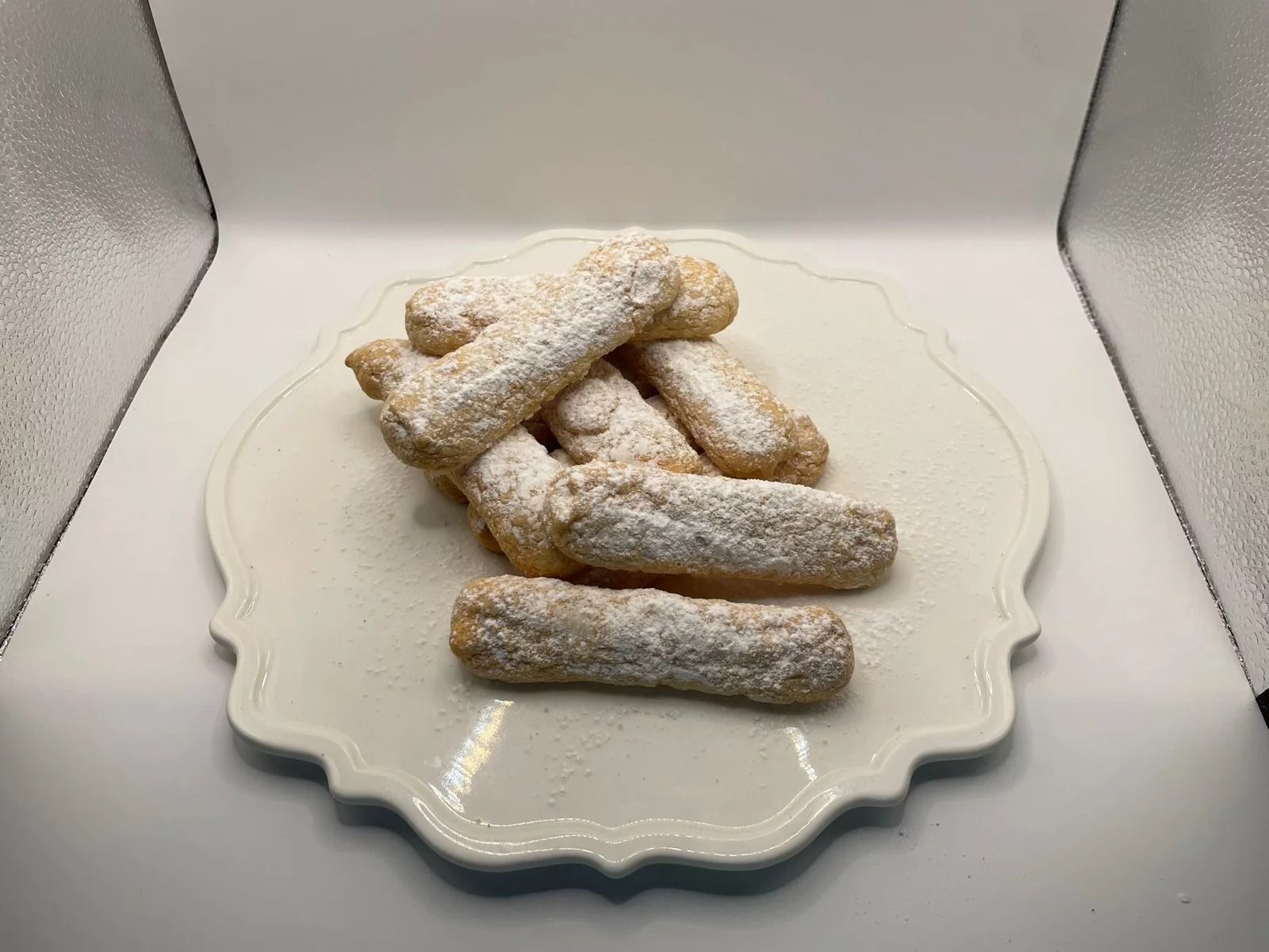 Mucchio di savoiardi ricoperti di zucchero a velo su un piatto bianco con i bordi smerlati.