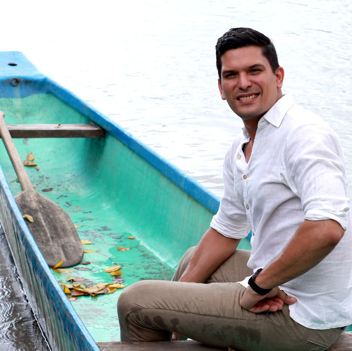 Hombre sonriente con camisa blanca sentado en un bote turquesa en aguas tranquilas.