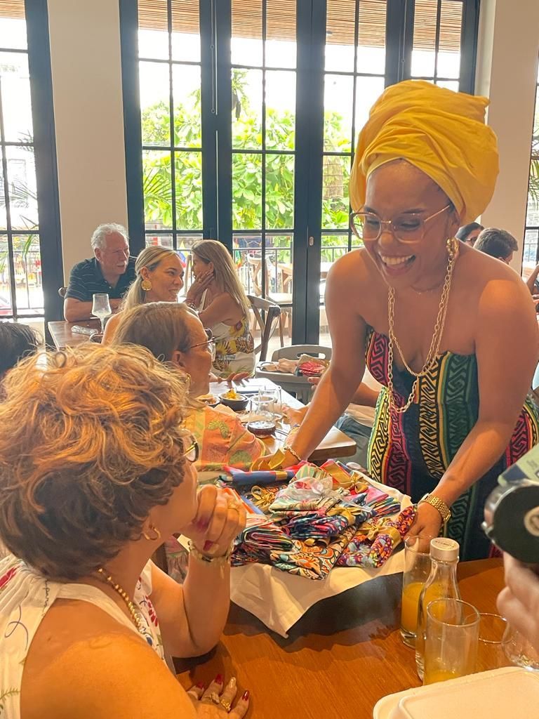 Una mujer con un turbante amarillo sirve un plato colorido en una mesa soleada con comensales sentados.