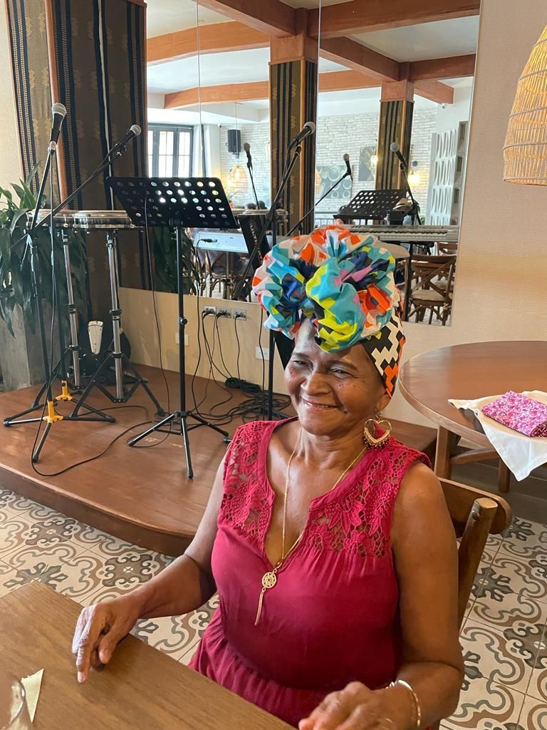 Una mujer sentada en el interior, sonriendo, con un vestido rojo y un pañuelo colorido en la cabeza