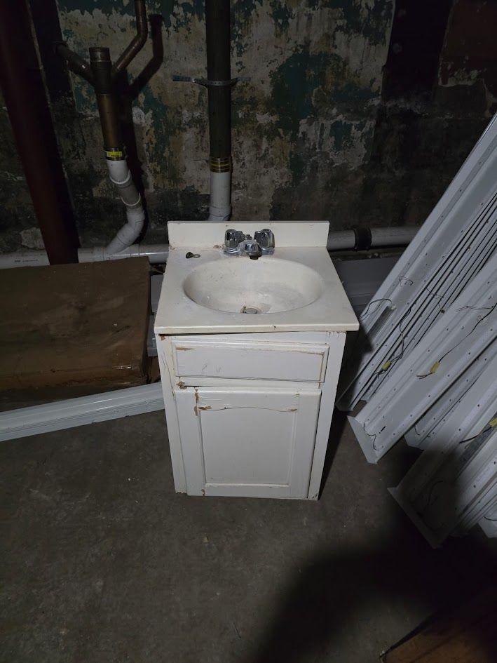 Small white bathroom vanity sink in a utility room with exposed pipes and stacked panels nearby