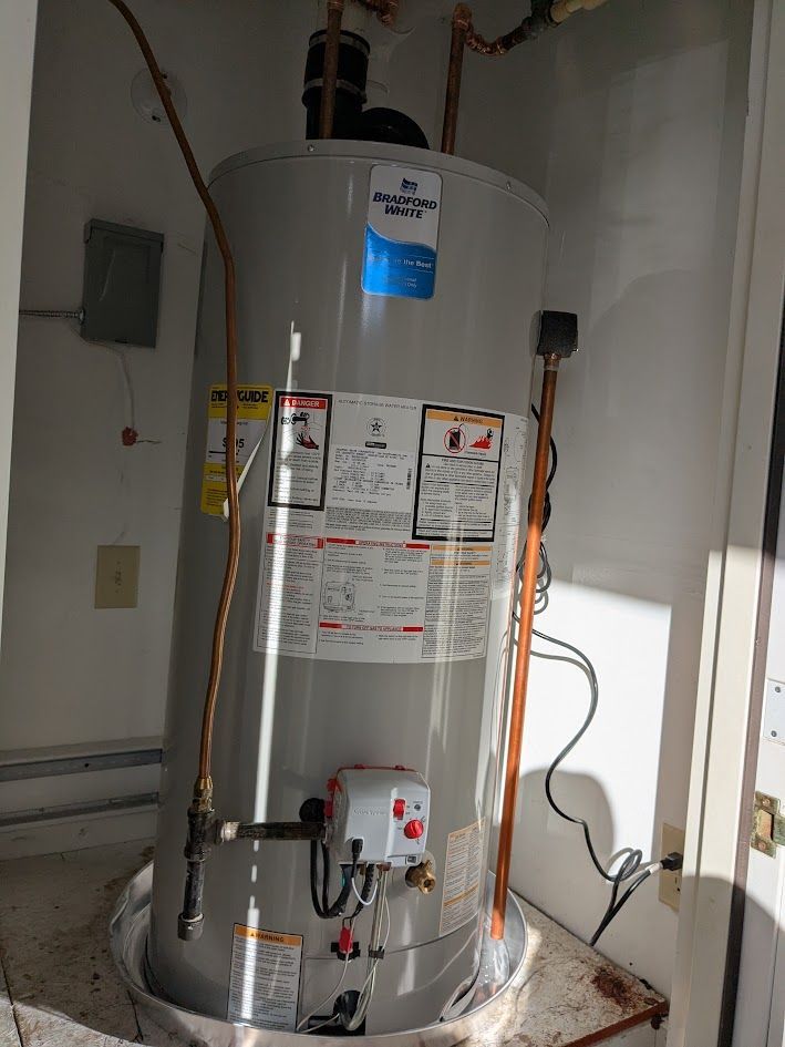 Tall silver water heater in a utility closet with warning labels and plumbing attachments