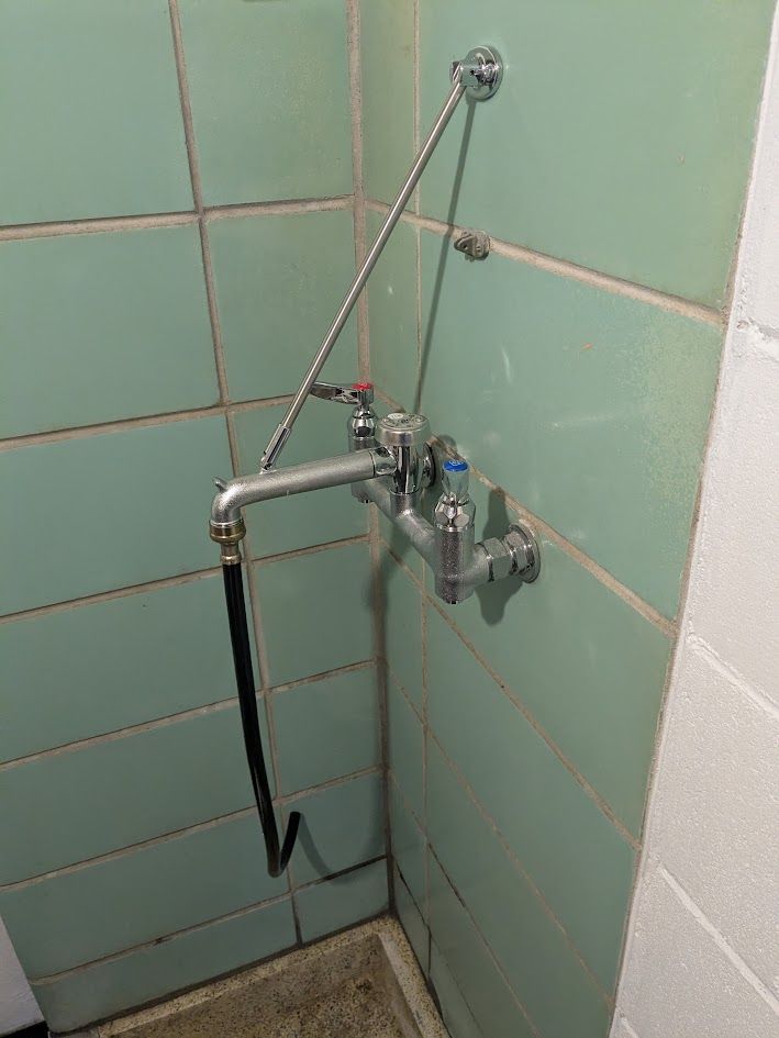 Wall-mounted shower hose and faucet in a tiled bathroom corner with a chain-operated drain stopper.