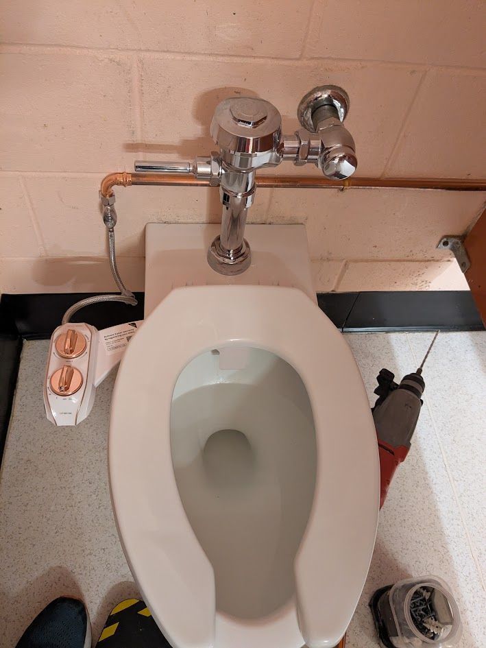 Public toilet with a wall-mounted flush valve and cleaning supplies on the floor.