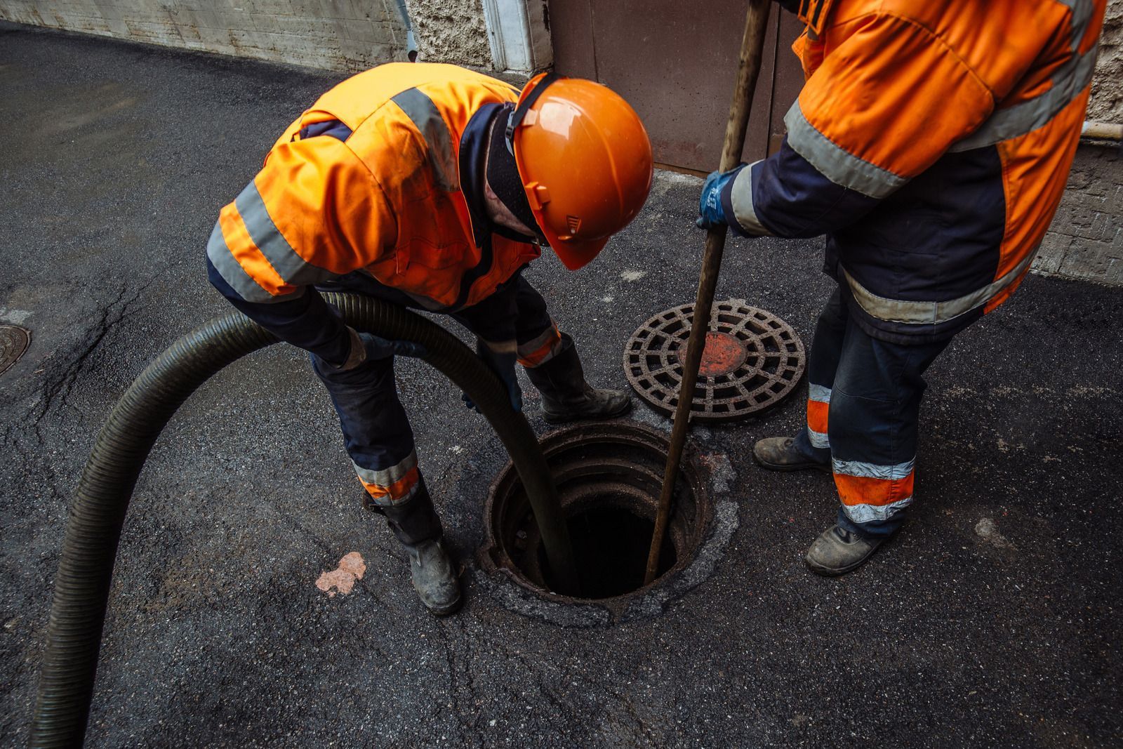 Two workers in orange safety gear inspecting a manhole on a wet road