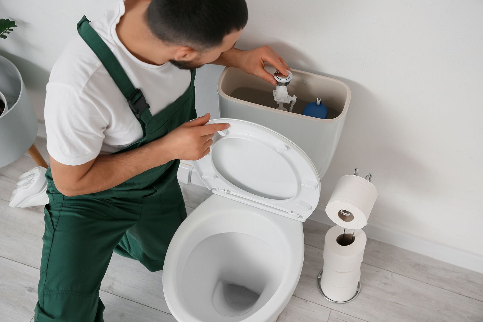 Technician in green overalls repairs a toilet’s tank in a bathroom.