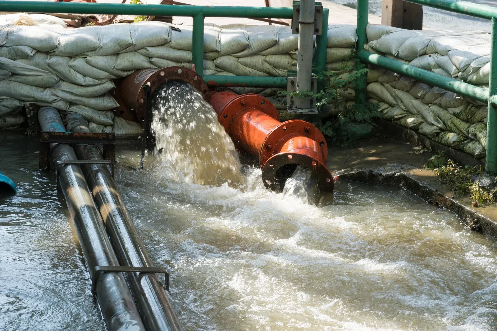 Water gushing from an orange pipe into a fenced containment area with muddy runoff and hoses.