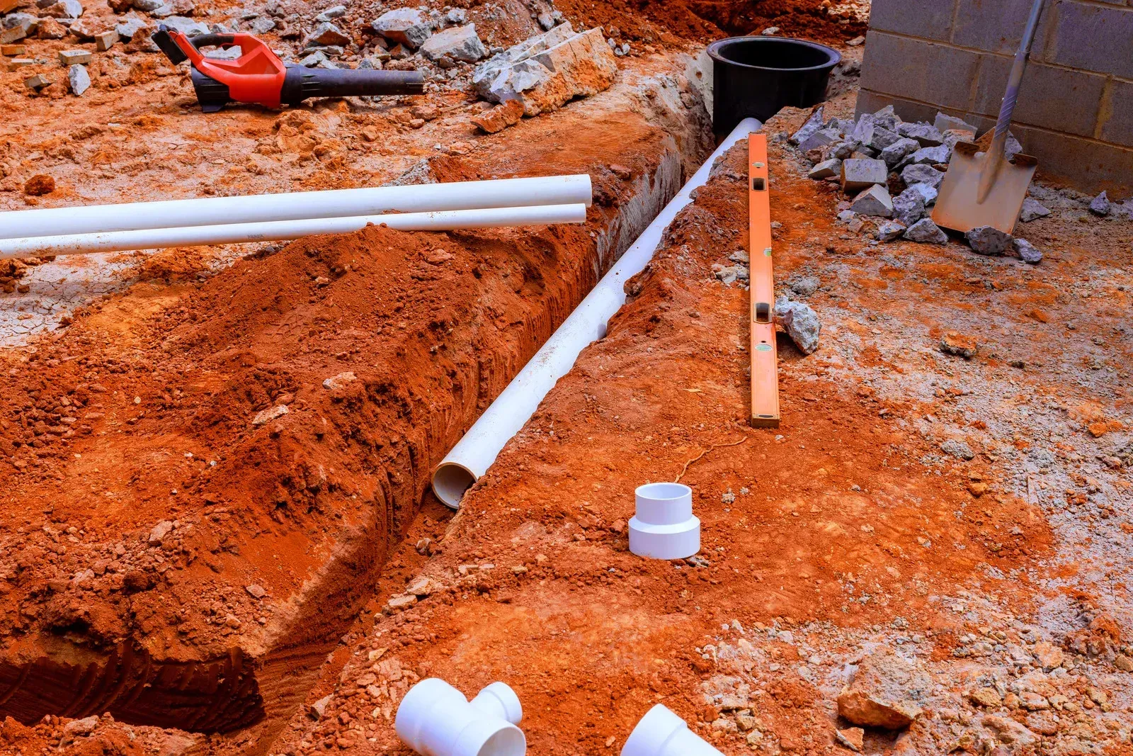 Trench with white PVC pipes and tools at a construction site with orange dirt and rubble