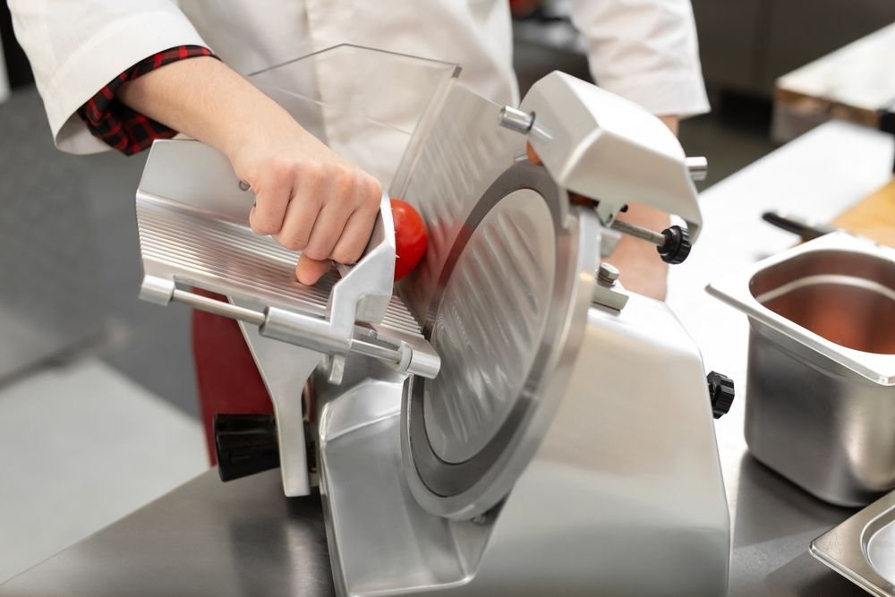 A person is cutting a tomato with a deli slicer.