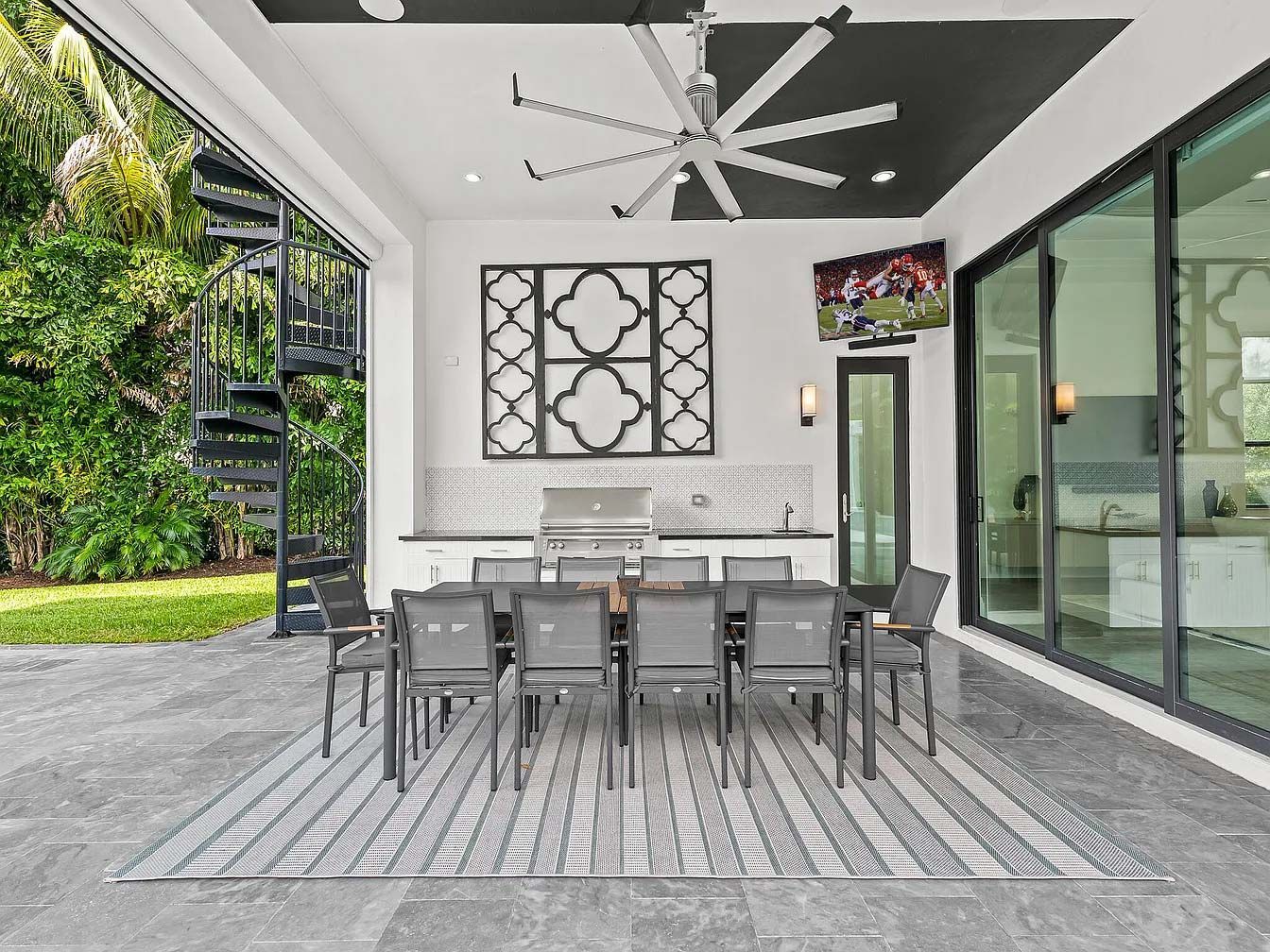 Covered outdoor dining patio with gray table and chairs, ceiling fan, and glass wall overlooking greenery