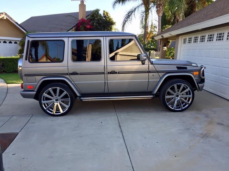 Silver Mercedes-Benz G-Wagon SUV with custom chrome wheels parked on a concrete driveway in front of a house.