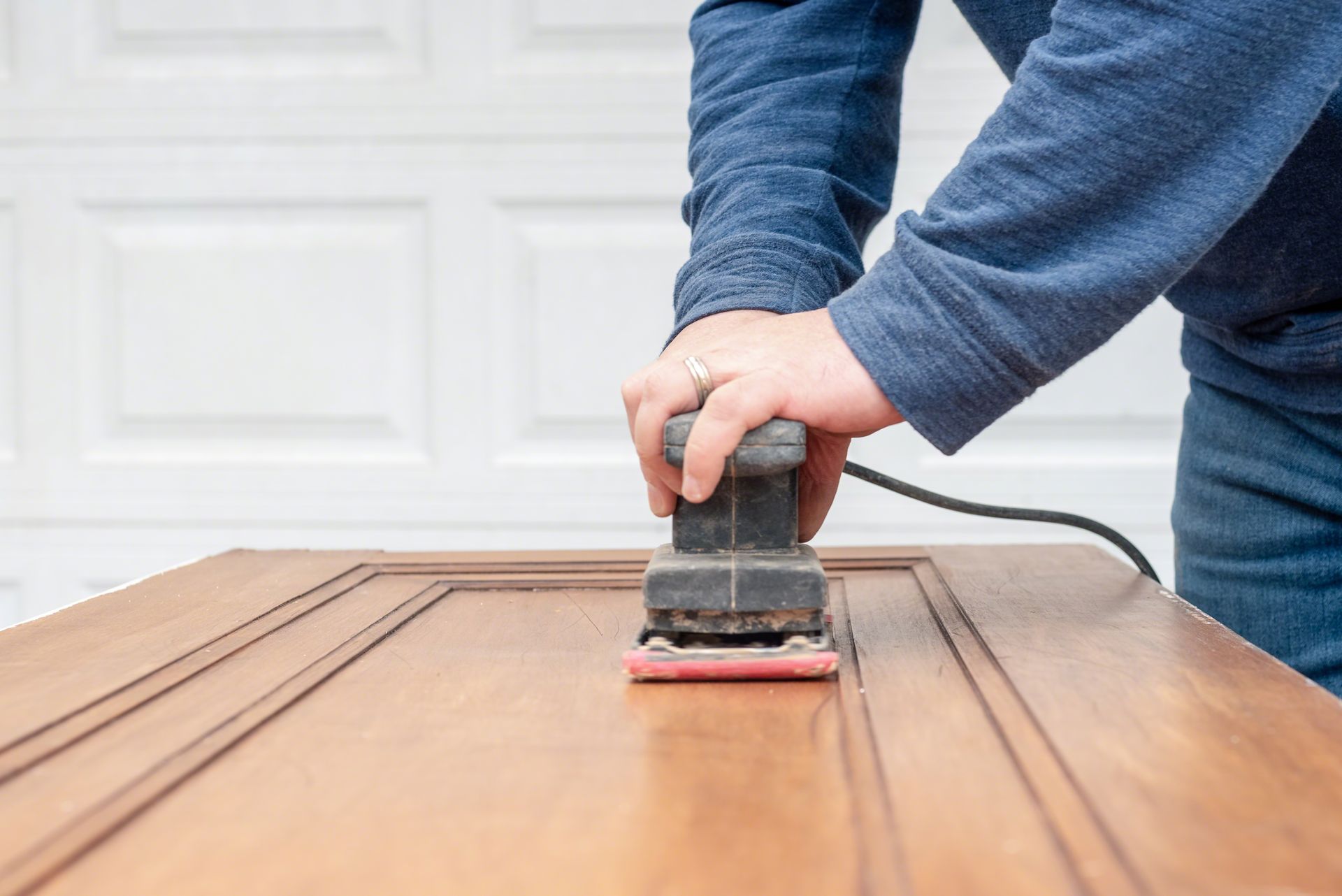A person uses a power sander to refinish a wooden cabinet door in a garage setting.