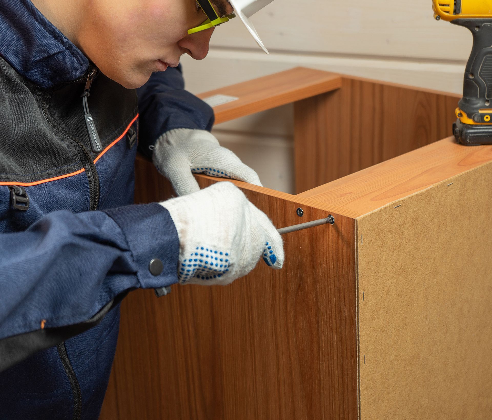 A person wearing a jacket and work gloves uses a manual screwdriver to secure a screw into wood cabinetry panels.