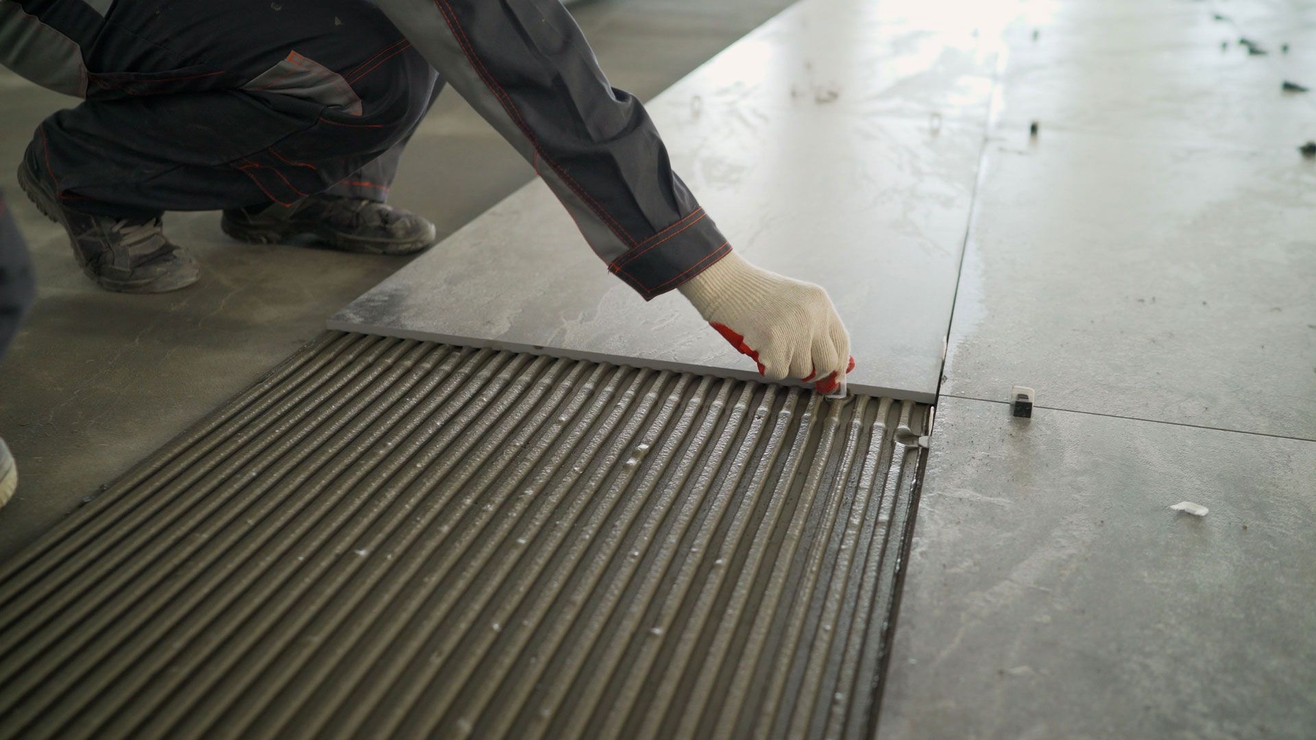 A worker laying a large tile onto a floor covered with ridges of fresh adhesive mortar.